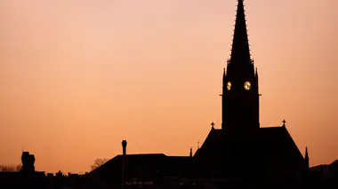 Silhouette d'un clocher d'église dont les horloges sont éclairées, sur un ciel orangé au coucher du soleil